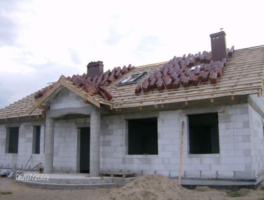 Budowa domu jednorodzinnego w trakcie realizacji: ściany z bloczków, dach z drewnianą konstrukcją i częściowo ułożoną dachówką ceramiczną, widoczne kominy i okno dachowe.